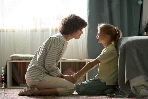 Young mother playing with her little daughter during her leisure time, they sitting on floor in bedroom and smiling to each other