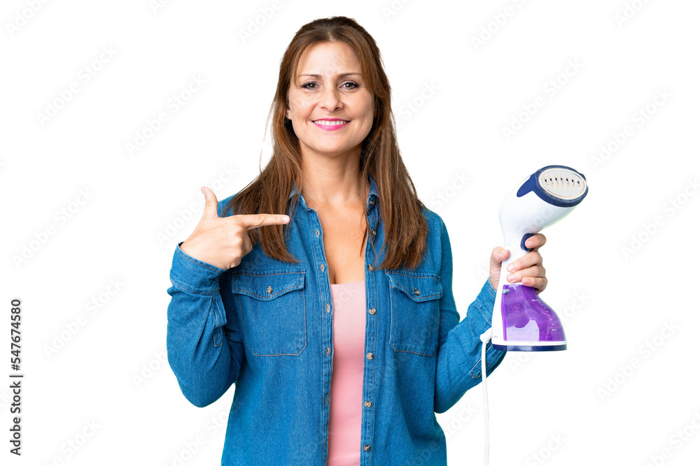Middle age woman holding an iron over isolated background and pointing it Middle age woman holding an iron over isolated background and pointing it