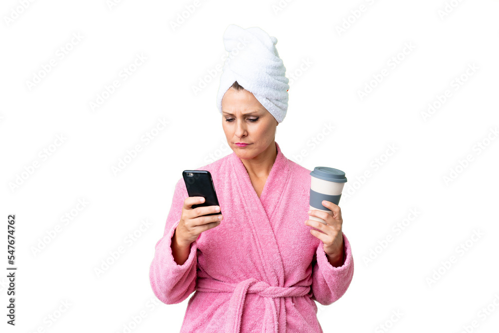 Middle age caucasian woman in a bathrobe over isolated background holding coffee to take away and a mobile