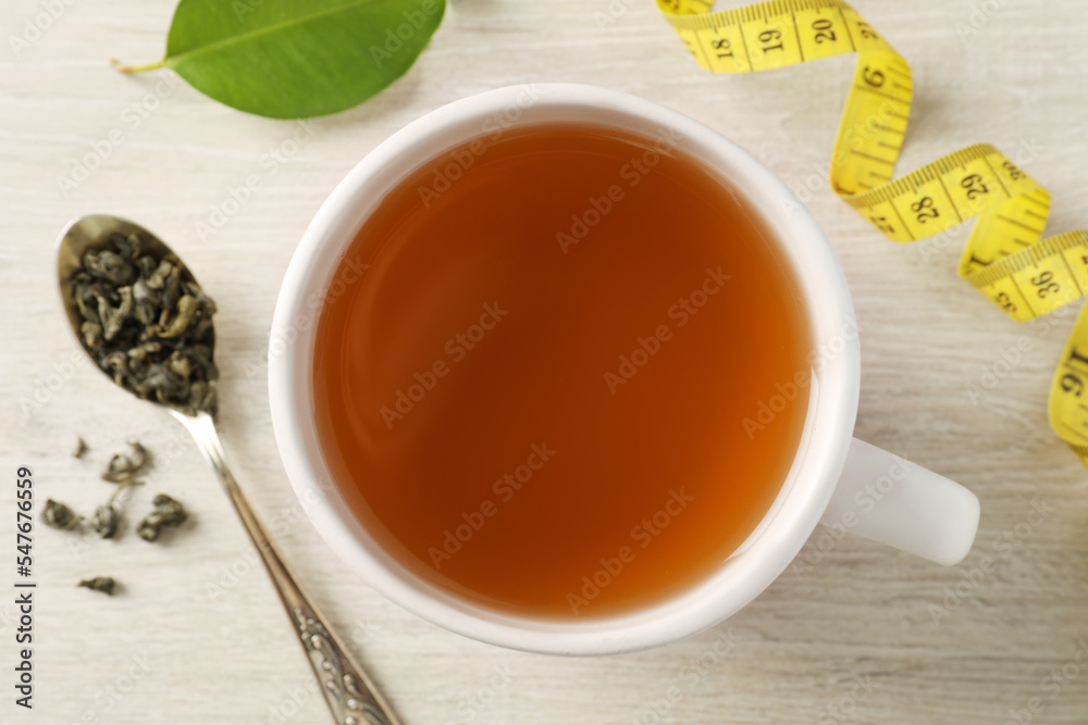 Cup of herbal diet tea and measuring tape on light wooden table, flat lay. Weight loss concept Cup of herbal diet tea and measuring tape on light wooden table, flat lay. Weight loss concept