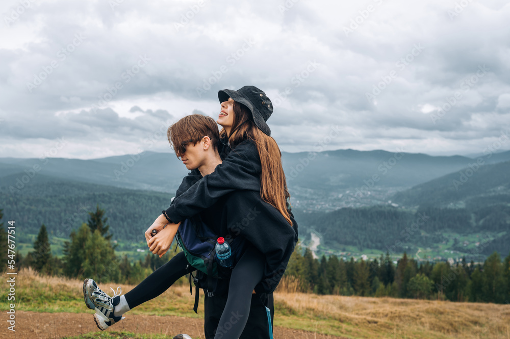 A young beautiful couple, a man and a woman, stand in the mountains on the background of a beautiful view, the man carries the woman on his shoulders.