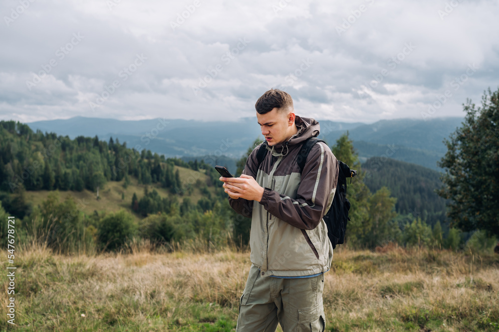 A serious male tourist on a hike stands in the mountains against the background of a beautiful landscape and uses a smartphone with a serious face.
