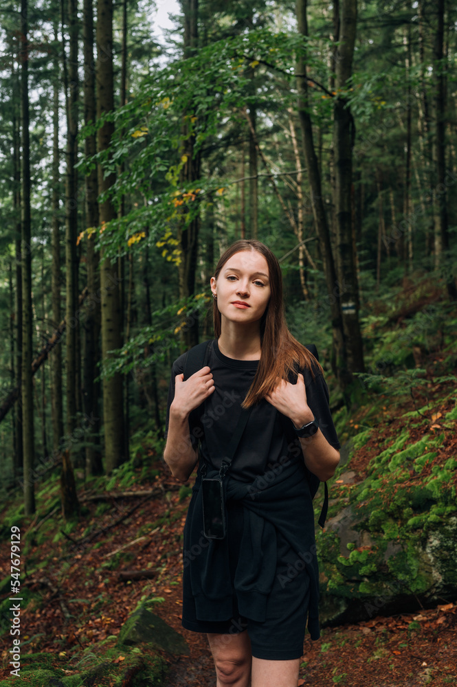 Portrait of an attractive female hiker in casual clothes standing on a path in the forest during a hike in the mountains and looking at the camera with a smile on her face. Portrait of an attractive female hiker in casual clothes standing on a path in the forest during a hike in the mountains and looking at the camera with a smile on her face.