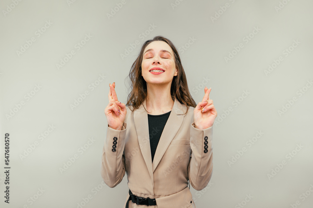 Cute woman in a suit makes a wish with her eyes closed, crossed her fingers and dreams with a smile on her face, isolated on a beige background. Cute woman in a suit makes a wish with her eyes closed, crossed her fingers and dreams with a smile on her face, isolated on a beige background.