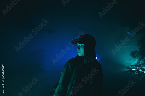 Night cinematic portrait of a woman in casual clothes and a cap looking away with a serious face on the street in the fog and in the green light.