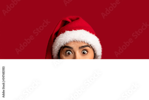Funny Arab Woman In Santa Hat Peeking Out Behind Blank Advertisement Board © Prostock-studio