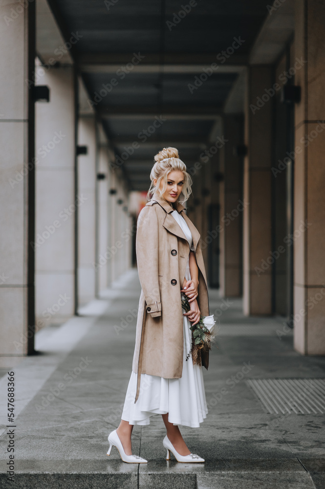 A beautiful blonde model bride in a beige coat stands in a city tunnel. Wedding photography, portrait, autumn.