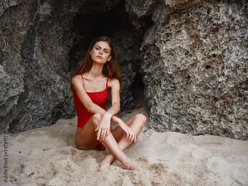 Woman in red swimsuit sitting by the rocks on the sand with a beautiful tan from the sun on the beach, travel to Bali, a tropical vacation