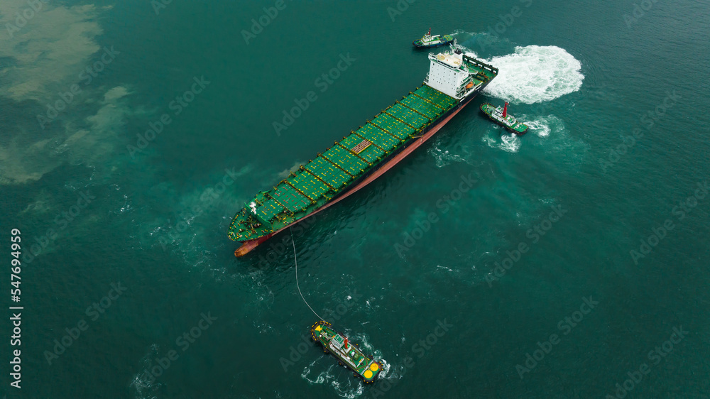 tugboat dragging cargo ship leaving shipyard after repairs in green sea, aerial top view