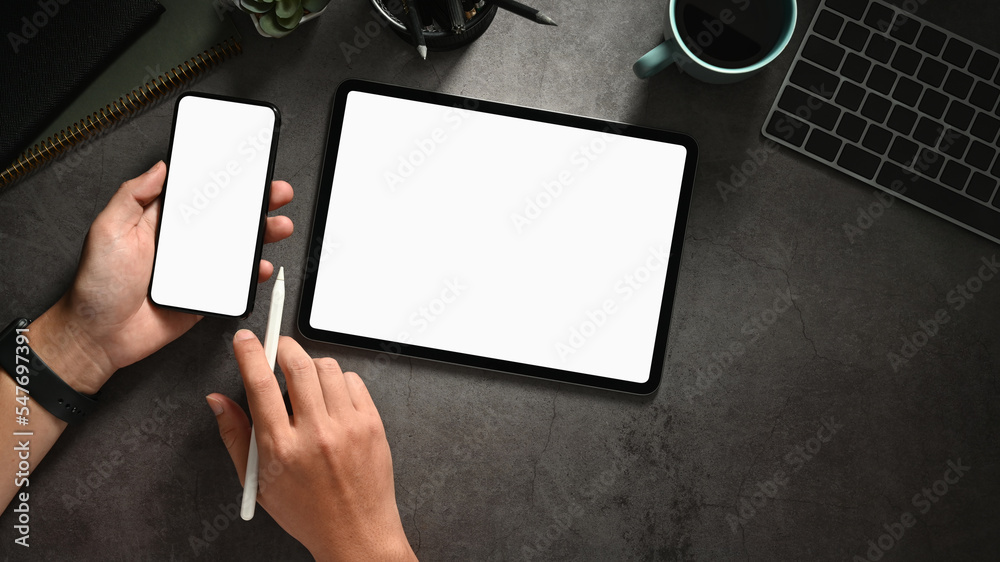 Obraz premium Top view office desk with hands holding smart phone and tablet, empty screen smartphone and tablet for your present.