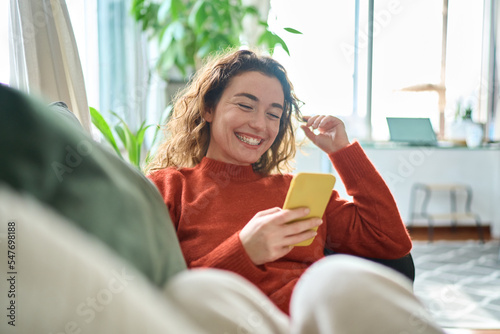 Happy relaxed young woman sitting on couch using cell phone, smiling lady laughing holding smartphone, looking at cellphone enjoying doing online ecommerce shopping in mobile apps or watching videos. photo