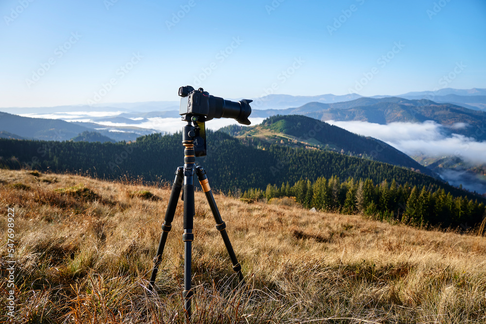 Camera on tripod on the meadow in the mountains capturing a beautiful mountain landscape with a fog, clouds and a forest. Landscape photography
