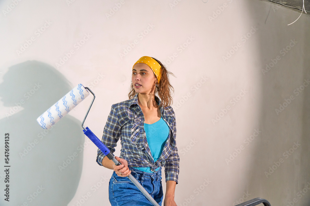 A young female artist paints the walls in the apartment with a roller. Beautiful woman is working on a new bedroom project. Cute woman in blue jeans and t-shirt painting a wall. Front view. A young female artist paints the walls in the apartment with a roller. Beautiful woman is working on a new bedroom project. Cute woman in blue jeans and t-shirt painting a wall. Front view.