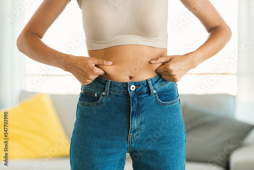 Young woman in jeans and white top squeezing her belly fat, planning training and eating healthy food