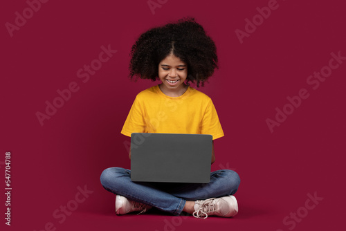 Cheerful black girl schooler using laptop, colorful background © Prostock-studio