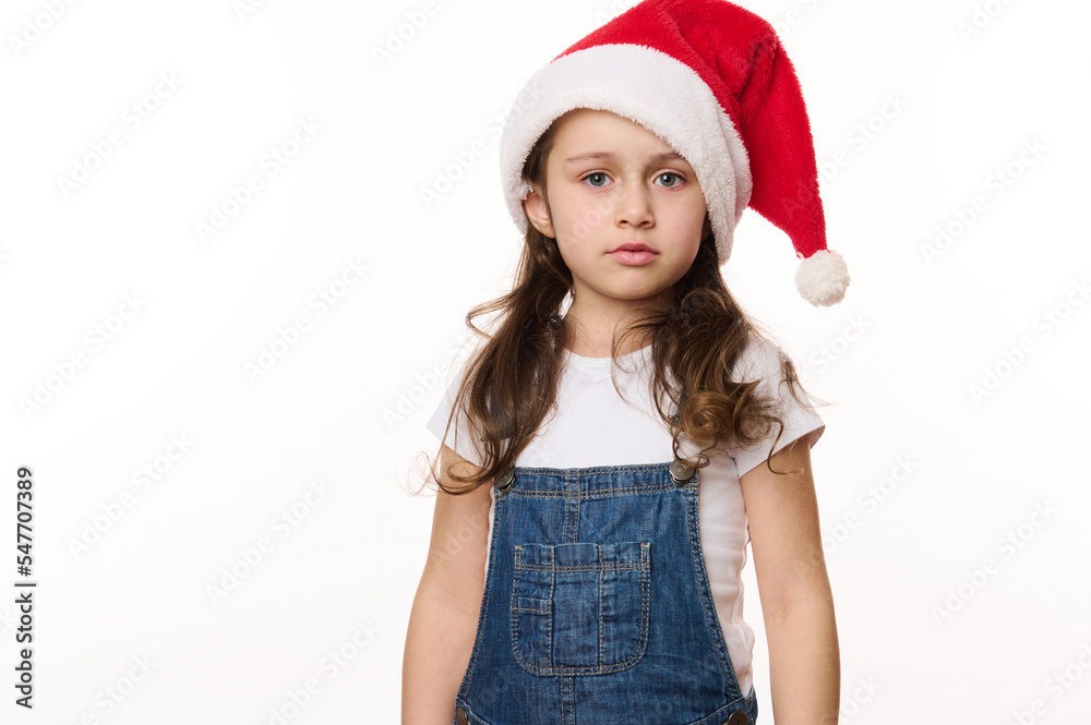 Isolated portrait on white background of a fancy cute Caucasian little girl in santa hat, in anticipation of Christmas holidays. Pure joy. Winter spirit. New Year party. Copy spacefor advertising text