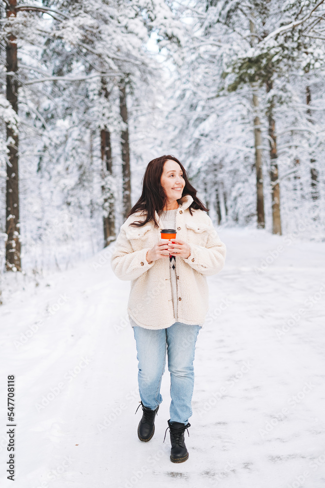 Smiling woman with dark hair in winter clothes with paper cup of coffee in hands against background of snowy winter forest