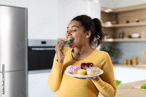 Smiling hungry young black pregnant female with big belly eats sweets, hold plate with cookies photo