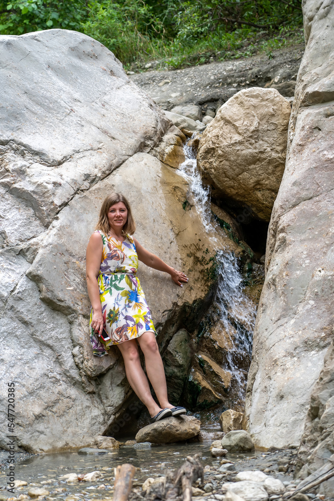 Carefree woman stands next to small stream flowing down from huge stones in mountain gorge onto pebbles Carefree woman stands next to small stream flowing down from huge stones in mountain gorge onto pebbles
