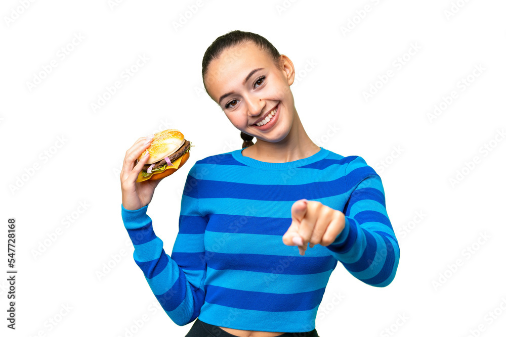 Young Arab woman holding a burger over isolated background pointing front with happy expression Young Arab woman holding a burger over isolated background pointing front with happy expression