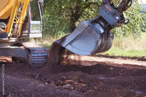 Erdbauarbeiten an einer Kabeltrasse auf dem Land. Der Bagger schaufelt mit seinem Baggerlöffel rotes Lockergestein. Sommer im Schwarzwald, Deutschland. 