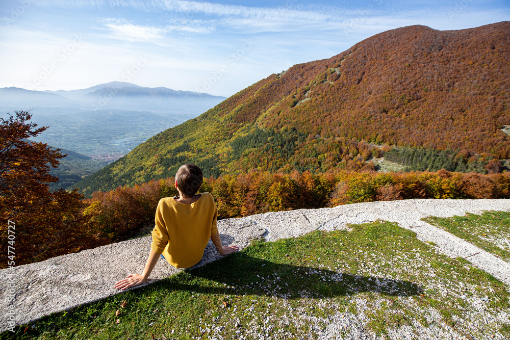 Fototapeta premium Young woman is sitting and looking at autumn landscape in Italy. Woman is sitting looking at the valley with its autumn colours.