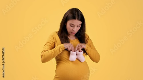 Happy prgnancy. Young playful pregnant lady playing with tiny booties, stepping on belly and laughing, yellow background photo