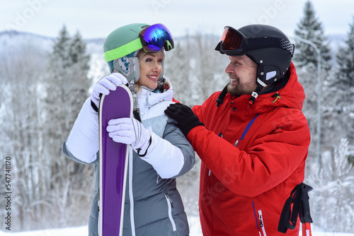 Happy loving couple smile at each other. They stand on the slope of a hill with mountain skis. Healthy lifestyle, winter outdoor activities, family relationships.