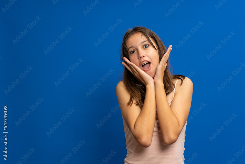 Pretty hispanic brunette teenage girl wearing a pink tank top and wearing a dental brace with her hands on her face making an adorable surprise gesture isolated over blue background. Pretty hispanic brunette teenage girl wearing a pink tank top and wearing a dental brace with her hands on her face making an adorable surprise gesture isolated over blue background.