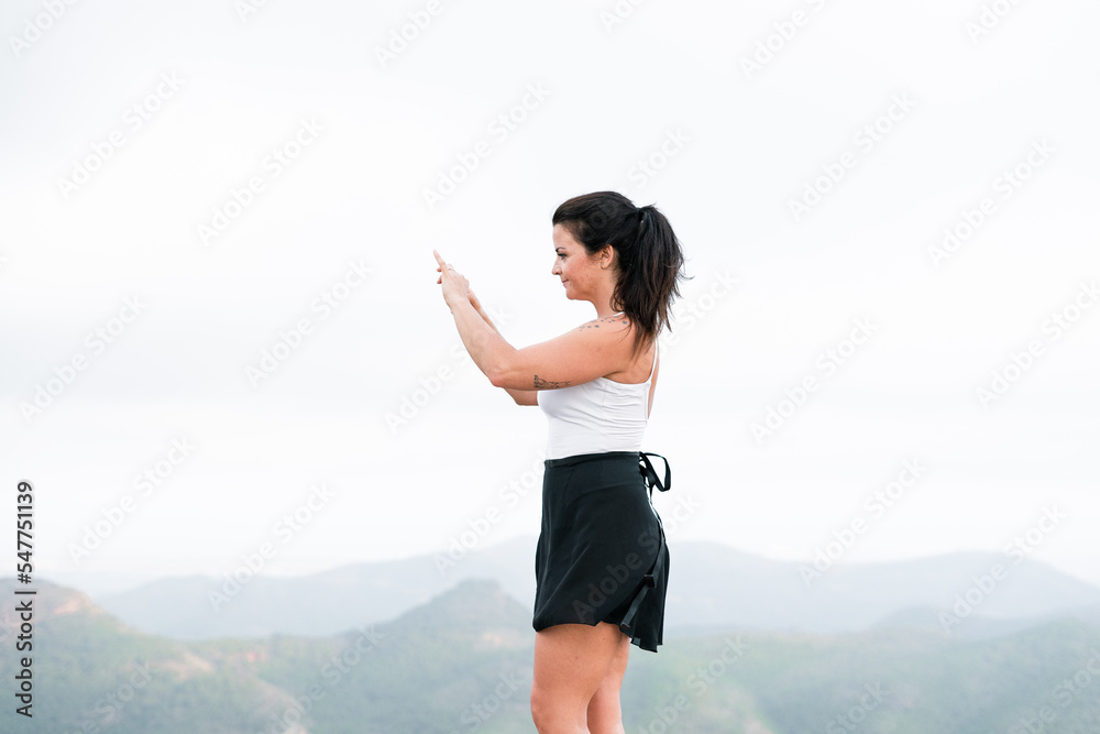 brunette caucasian mature woman in white top and black skirt making a photo of herself with her smartphone on top of mountain brunette caucasian mature woman in white top and black skirt making a photo of herself with her smartphone on top of mountain