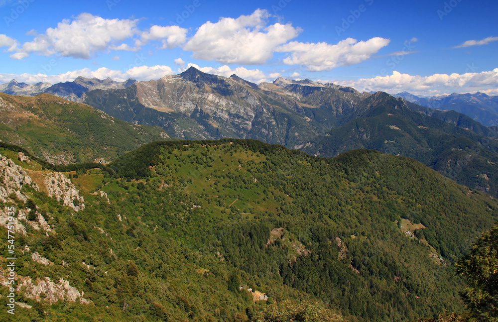 Panoramic view of the landscape with mountains and green hills in the foreground against a blue sky with clouds on Mount Cimetta, near Locarno in Switzerland