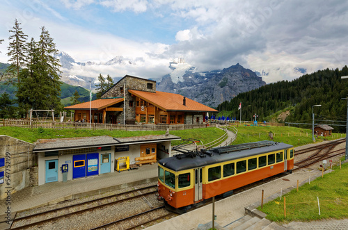 Scenery of a local train arriving at Winteregg Station near Murren in Bernese Oberland, Switzerland, with a restaurant on the grassy meadow and Jungfrau Mountain hiding in the clouds in background