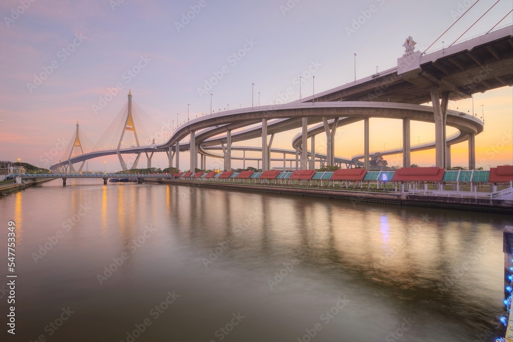 Low angle view of Bhumibol Suspension Bridge (or Industrial Ring Road ) over Chao Phraya River under evening twilight in Bangkok Thailand, with beautiful reflections on smooth water at red rosy dusk