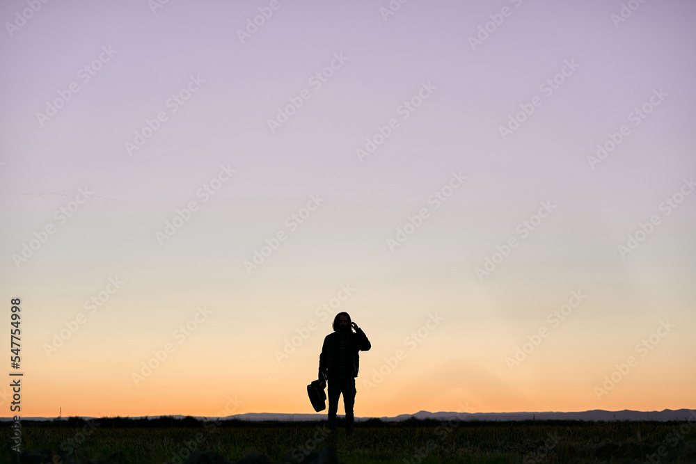 silhouette of a caucasian boy with long hair standing on the green plain calm and relaxed while holding a guitar