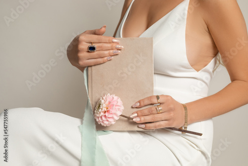 Unrecognizable cropped woman in white dress with decollete holding blank diary notebook and flower in hands in studio