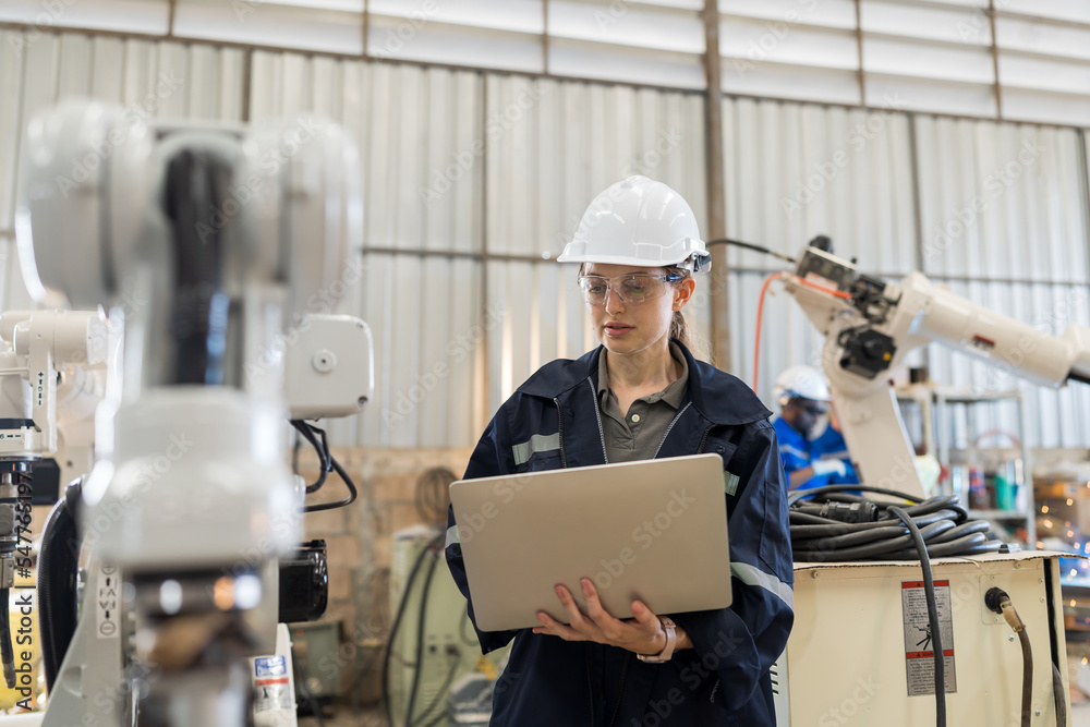 Female engineers worker using laptop computer working with adept robot arm in workshop. Female technician maintenance automatic robot arm system welding with laptop computer in factory Female engineers worker using laptop computer working with adept robot arm in workshop. Female technician maintenance automatic robot arm system welding with laptop computer in factory