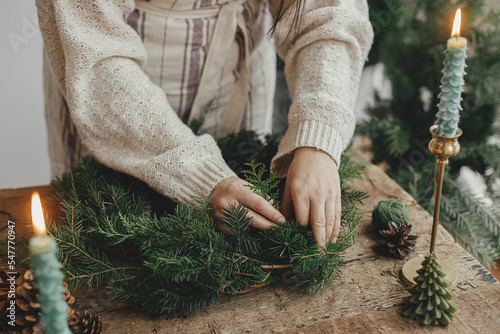 Making Christmas rustic wreath. Woman hands holding fir branches and making wreath on rustic wooden table with ribbon, golden bells, candles. Moody holiday image. Winter holiday workshop