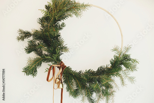 Modern Christmas wreath with velvet ribbon and golden bells hanging on white wall background close up. Merry Christmas and Happy Holidays! Winter holiday decor, moody image. Boho wreath photo