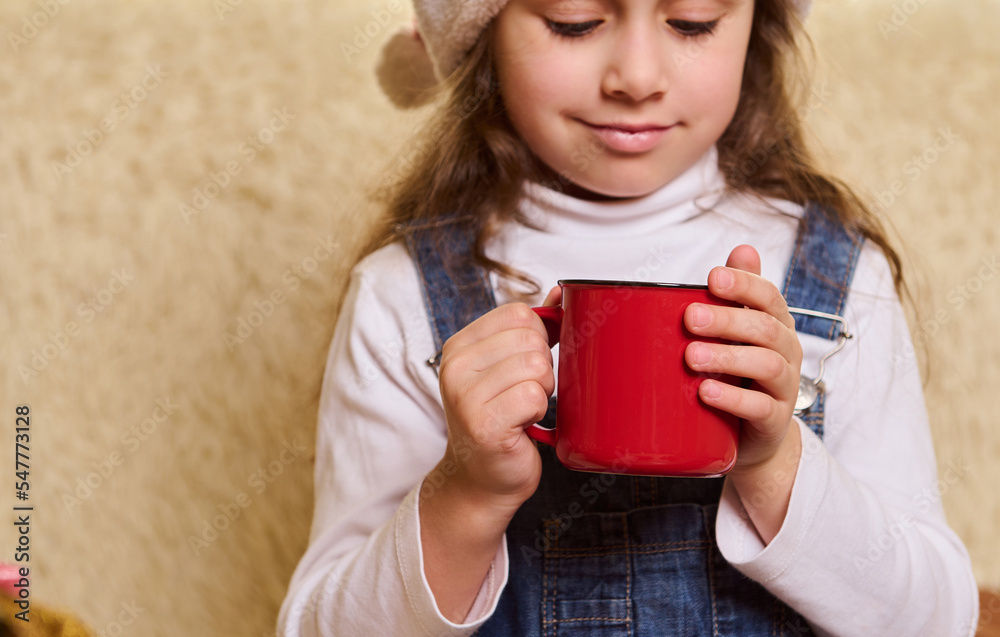 Details: red cup of hot chocolate or cocoa drink with marshmallows in the hands of a cute baby girl, wearing Santa hat, enjoying happy festive atmosphere of Christmas winter holidays at home interior