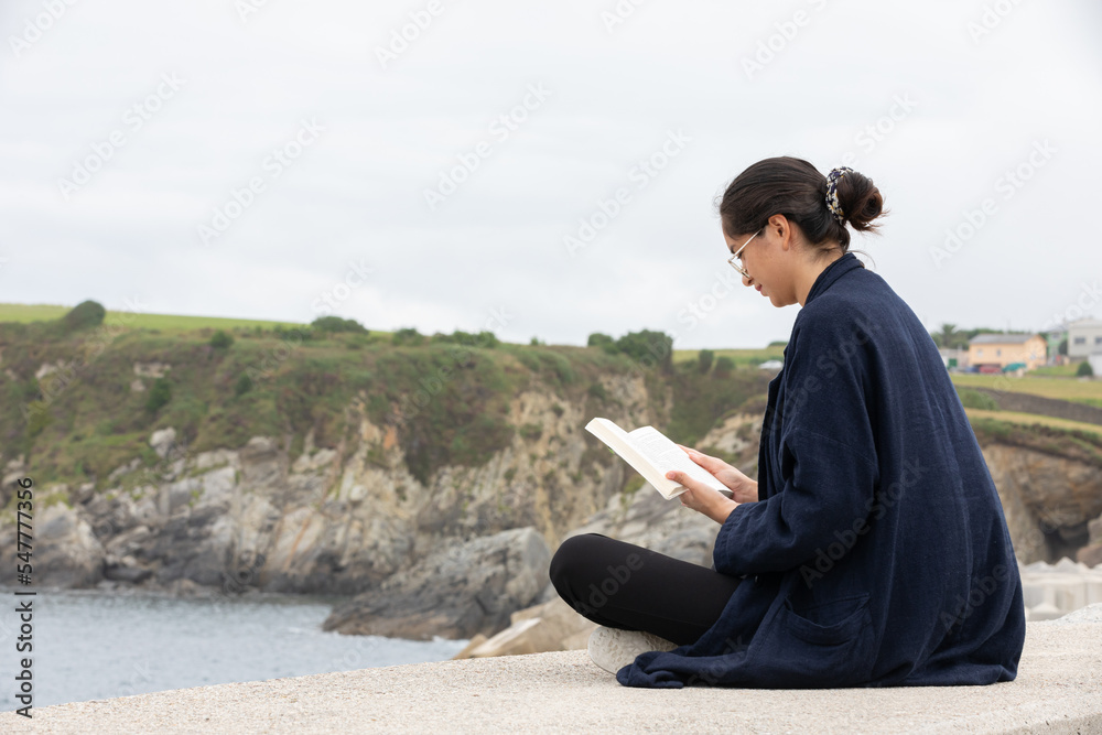 Side view of female in casual clothes reading book near sea 