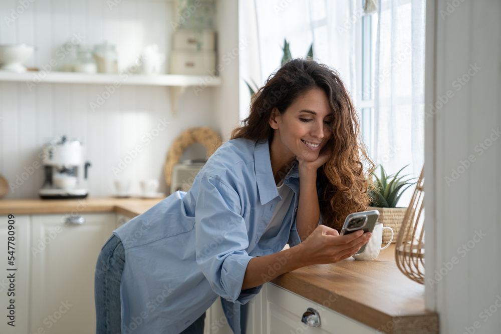 Happy pleasant young woman using smartphone, enjoying online shopping, spending leisure time at cozy home. Smiling female standing in kitchen holding mobile, surfing internet or chatting with friends