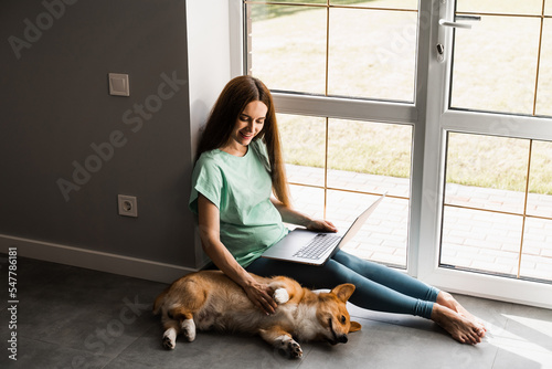 Pregnant girl with laptop smile and play with Corgi dog at home. Programmer woman working online and have a break for hug her dog. Having fun with Welsh Corgi Pembroke.