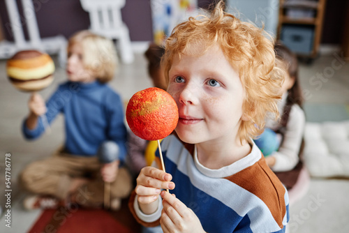 Adorable gingerhaired boy holding planet model of Mars by his face while studying astronomy at lesson in nursery school