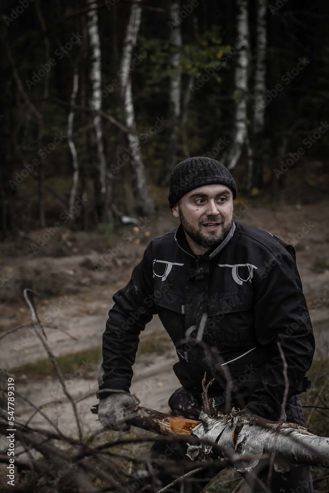 A man with a beard pulls a tree trunk with branches from a pile of fallen trees. A man in work clothes sorts a blockage of trees. The concept of a strong man. A man with a beard pulls a tree trunk with branches from a pile of fallen trees. A man in work clothes sorts a blockage of trees. The concept of a strong man.