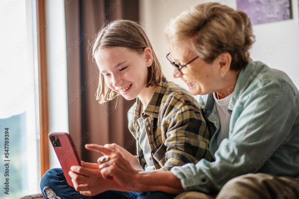 Smiling girl granddaughter with happy grandmother having fun and using looking at smartphone screen