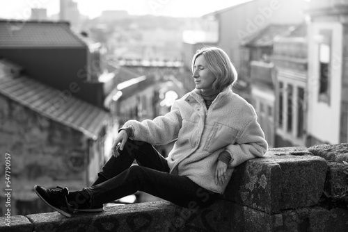 A female tourist sits on a masonry wall in Porto, Portugal. The houses in the background are blurred. Black and white photo.