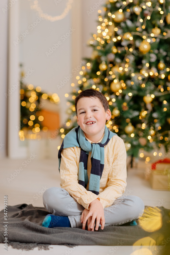 Little boy sits in front of the fir tree. Beautiful Christmas backdrop. Christmas mood at home. Child rejoices in Christmas and gifts.