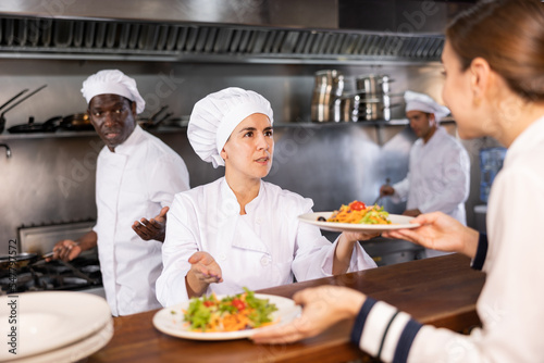 Portrait of smiling young woman chef working in restaurant kitchen  giving out ready salads s to waitress on order station