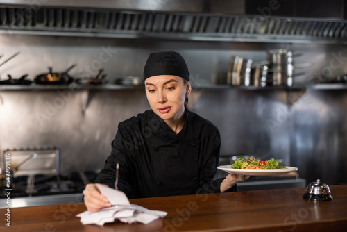 Portrait of skilled young woman chef giving out finished dish at ordering station and reading new orders in restaurant kitchen