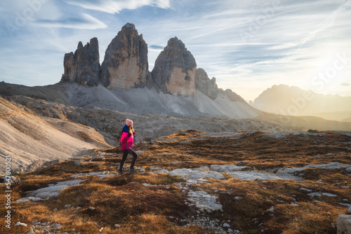 Walking girl with backpack on the trail in mountains at sunset in autumn. Tre Cime, Dolomites, Italy. Beautiful landscape with young woman, high rocks, path, stones, orange grass, sky in fall. Hiking